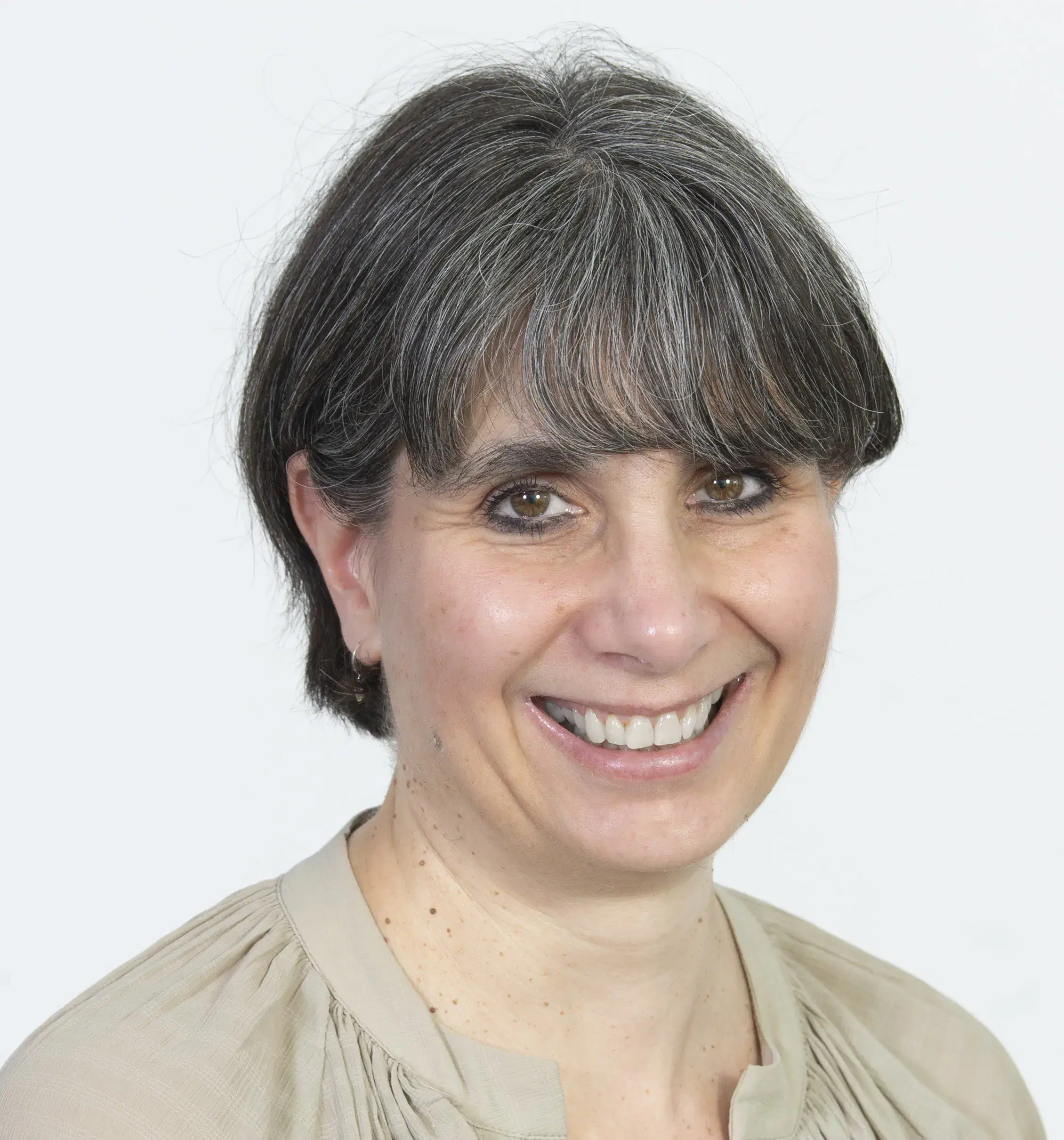 A woman with short, grey-streaked hair and brown eyes smiles at the camera. She is wearing a beige top and small hoop earrings against a plain white background.