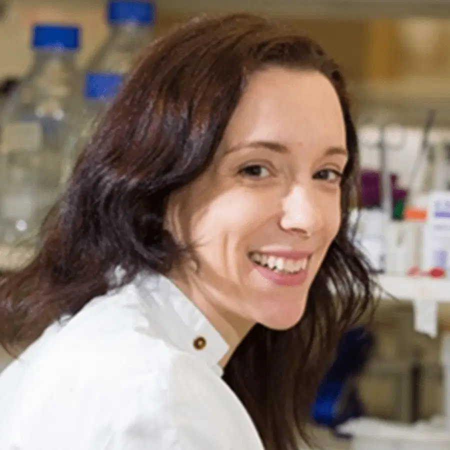 A woman in a white lab coat smiles whilst sitting in a laboratory with shelves of bottles and scientific equipment visible in the background.