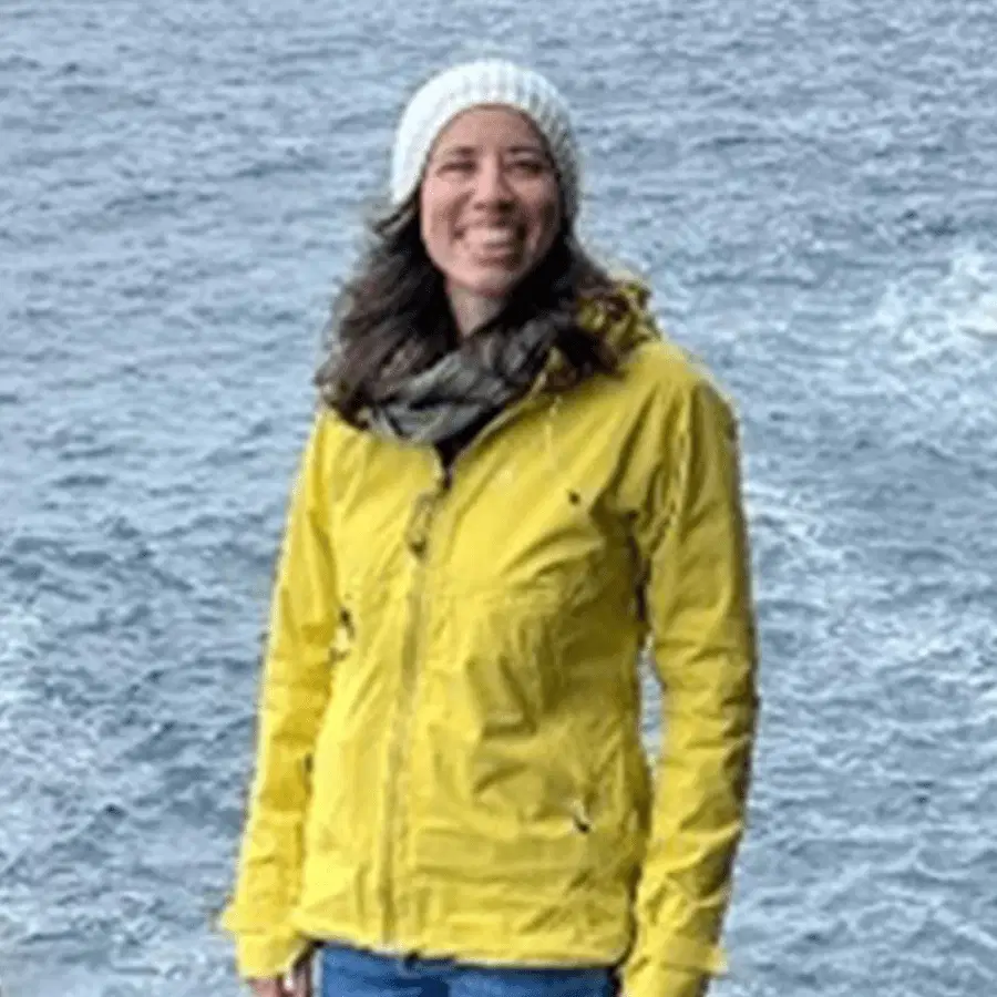 A smiling person in a yellow jacket and white knitted hat stands near a body of water with waves visible in the background.