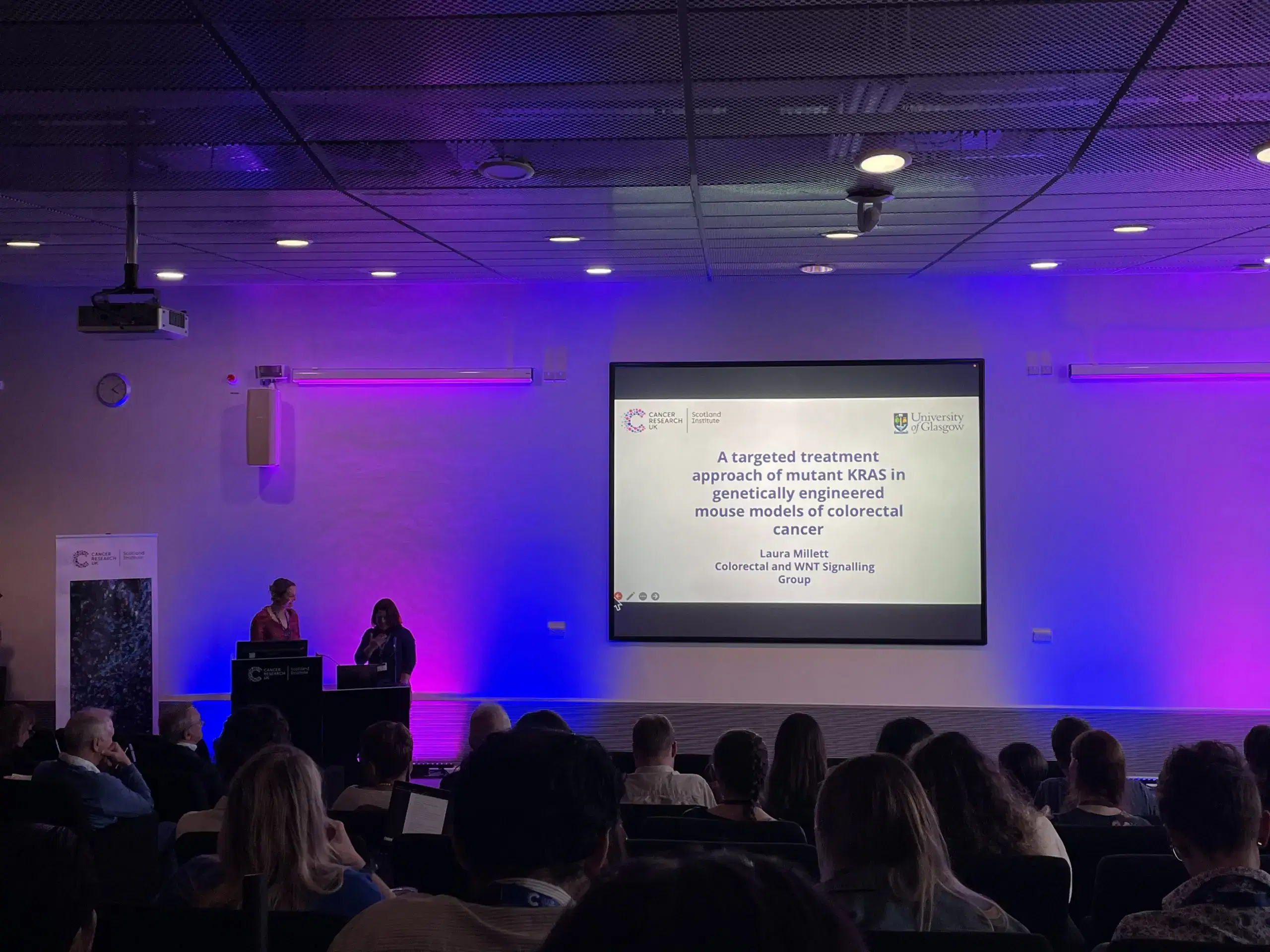 Audience watching a scientific presentation titled A targeted treatment approach of mutant KRAS in genetically engineered mouse models of colorectal cancer on a projected screen in a dimly lit conference room.