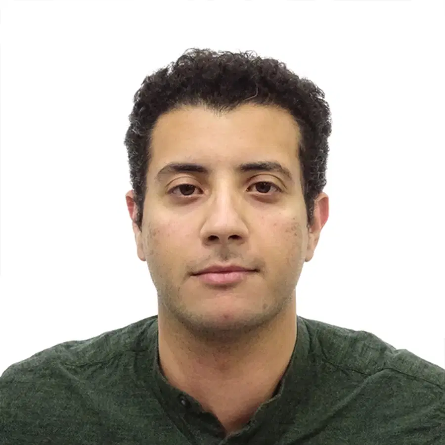 A man with short, curly dark hair and medium skin tone wears a dark green buttoned shirt, facing the camera with a neutral expression against a plain white background.