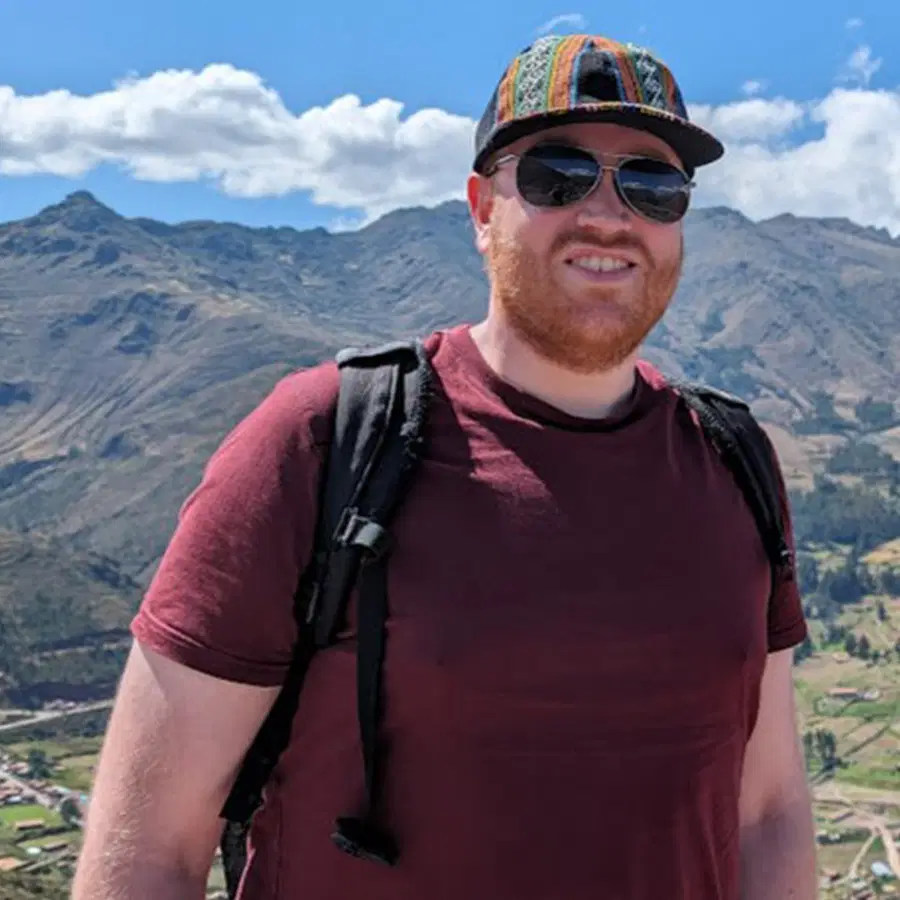 A man with a beard wearing sunglasses, a colourful cap, a maroon shirt, and a black rucksack smiles outdoors with mountains and a blue sky in the background.