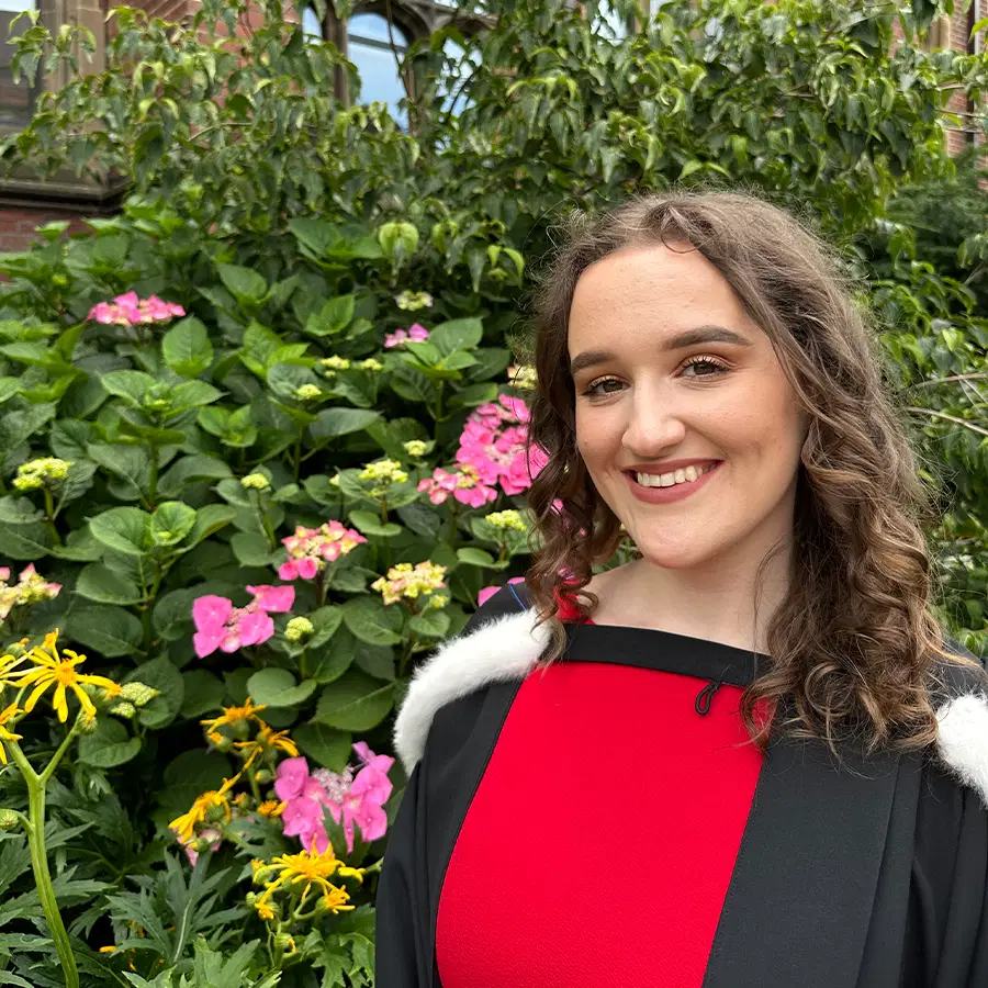 A young woman in a graduation gown and red dress smiles in front of green shrubs with pink and yellow flowers.