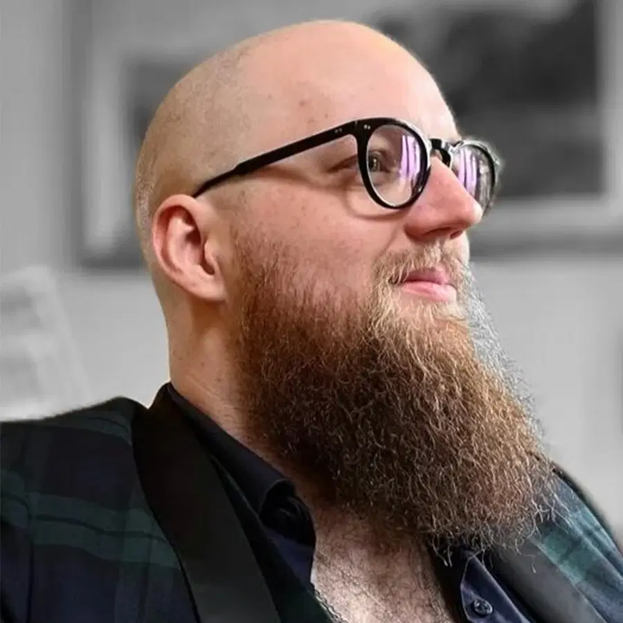 A bearded man with glasses and a shaven head looks to the side, smiling slightly. He is wearing a checked jacket and is indoors, with a blurred background.