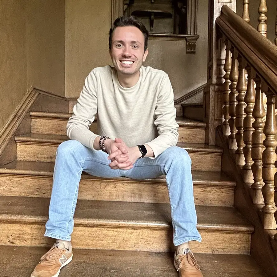 A smiling person in a beige jumper and light blue jeans sits on wooden stairs with hands clasped, next to a banister, indoors.