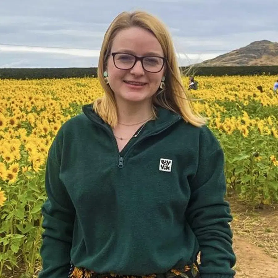 A person with glasses and a green jumper stands in a sunflower field, smiling at the camera. Hills and a cloudy sky are visible in the background.