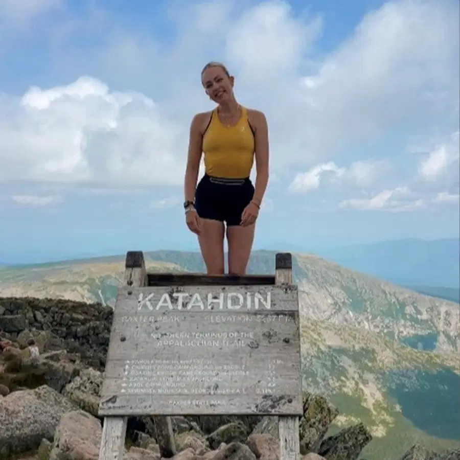 A woman in a yellow top and black shorts smiles, standing behind a Katahdin summit sign on a rocky mountaintop with mountains and blue sky in the background.