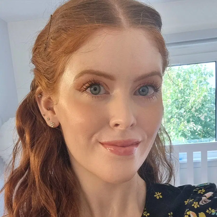 A woman with long wavy red hair smiles at the camera in a bright room. She is wearing a black floral top and has subtle make-up. A window with green trees outside is visible in the background.