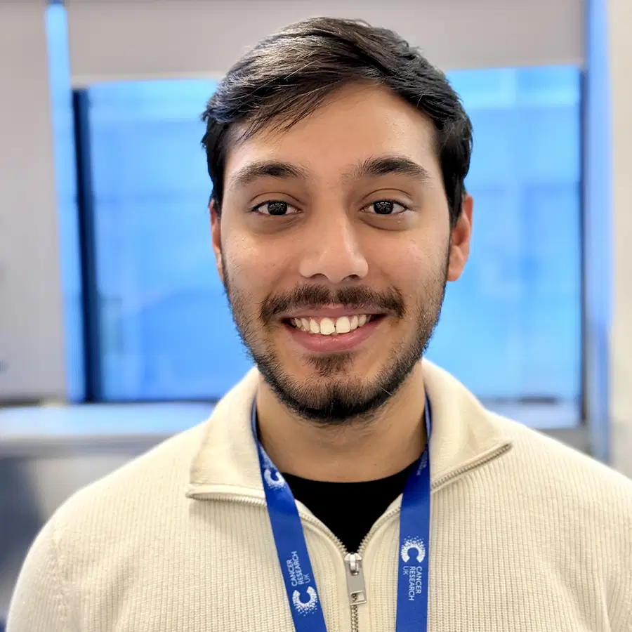 A man with short brown hair and a beard smiles at the camera. He wears a beige zip-up jumper and a blue lanyard. The background is an indoor setting with blurred windows.