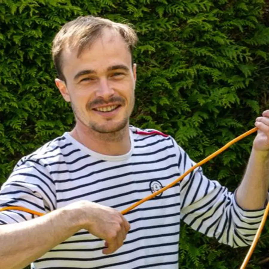 A smiling man in a white and black striped shirt holds an orange extension lead outdoors, with green leafy bushes in the background.