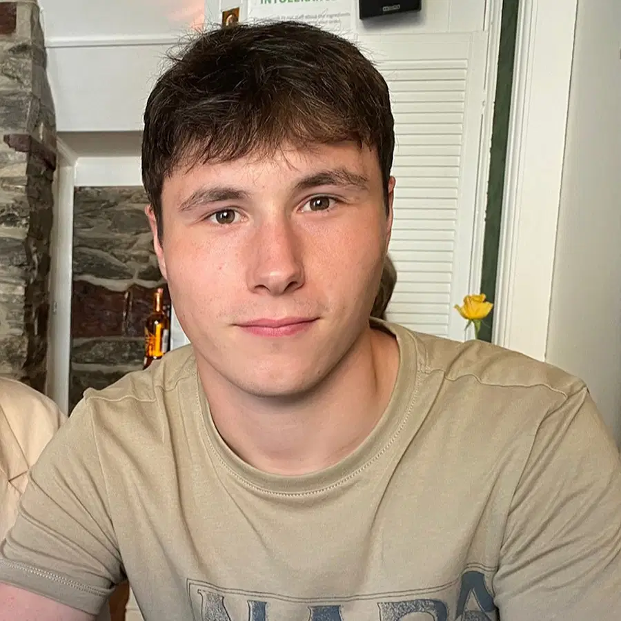 A young man with short brown hair wearing a beige T-shirt sits indoors, looking directly at the camera. The background shows a white door, a stone wall, and some decor items.