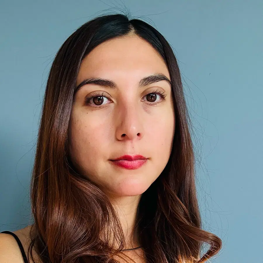 Woman with long brown hair and red lipstick poses against a plain blue background, looking directly at the camera with a neutral expression.