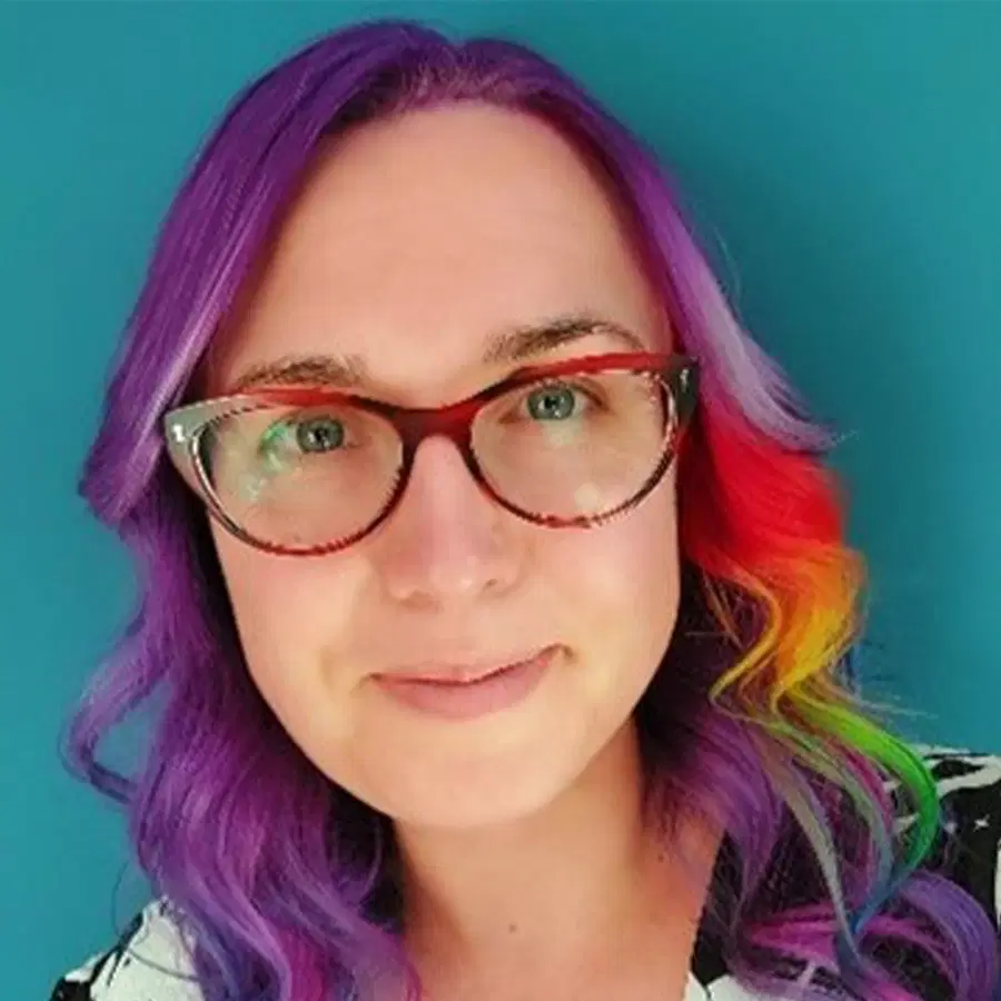 Person with long purple hair and a rainbow streak wears red-framed glasses and a patterned shirt, smiling softly against a solid blue background.