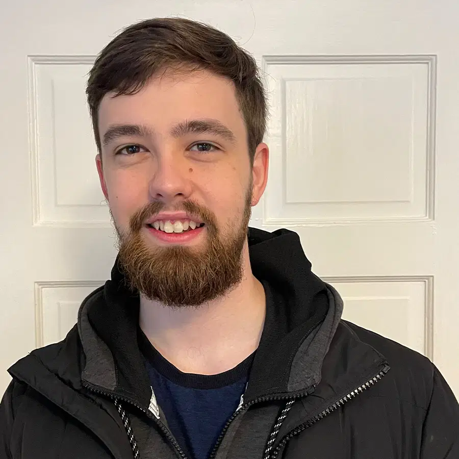 A young man with short brown hair and a beard smiles whilst wearing a black jacket and hoodie, standing in front of a white panelled door.