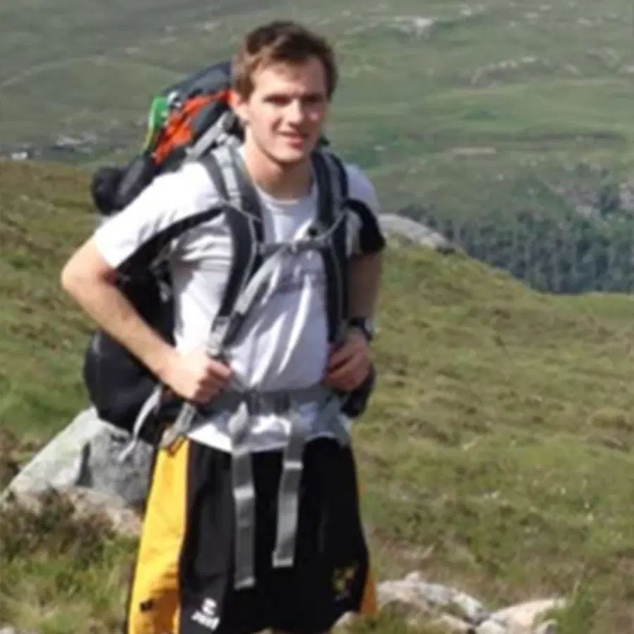 A person wearing a rucksack and outdoor kit stands on a grassy hillside, with rolling green hills and rocks in the background.