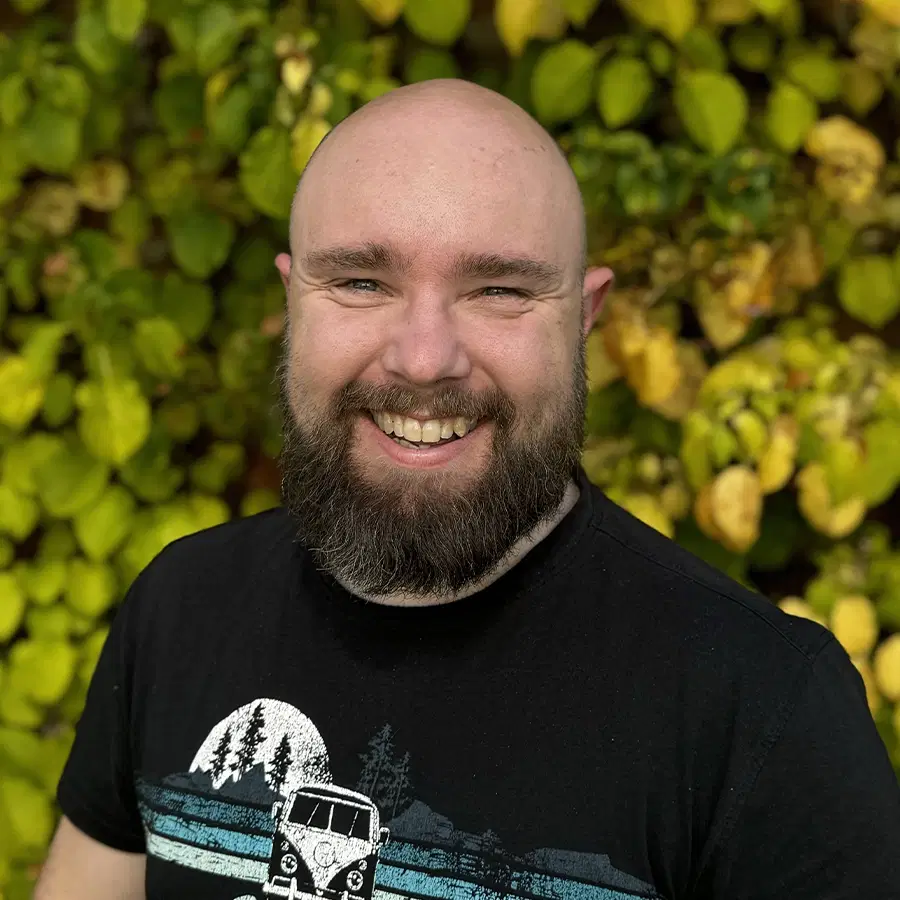 A bearded man with a shaved head smiles whilst standing in front of a wall covered in green and yellow leaves. He wears a black T-shirt with a graphic design on it.