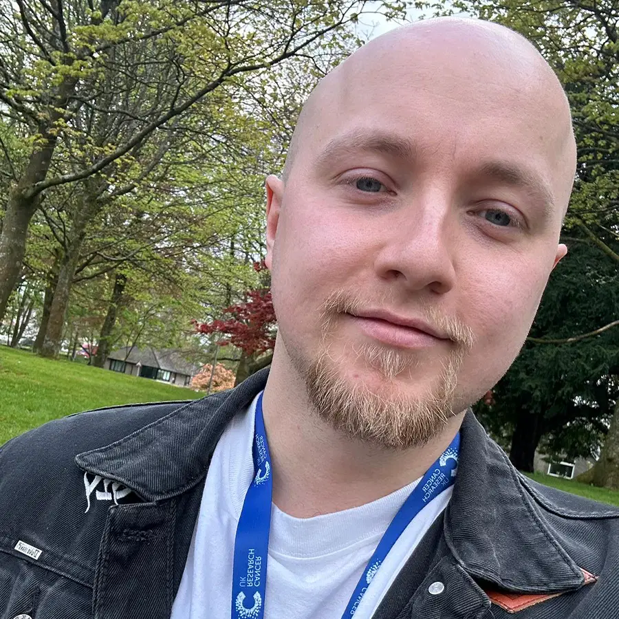 A person with a goatee and a blue lanyard smiles outdoors in a park, wearing a black jacket and white shirt. Green grass, trees, and overcast sky are visible in the background.
