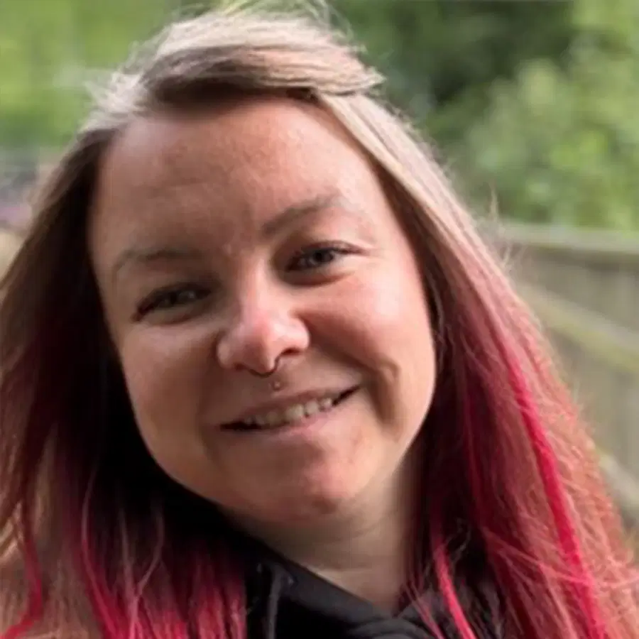 A person with long, dark hair featuring pink tips smiles outdoors. They have light skin, a nose ring, and are wearing a dark top. Green trees and a wooden railing are visible in the blurred background.
