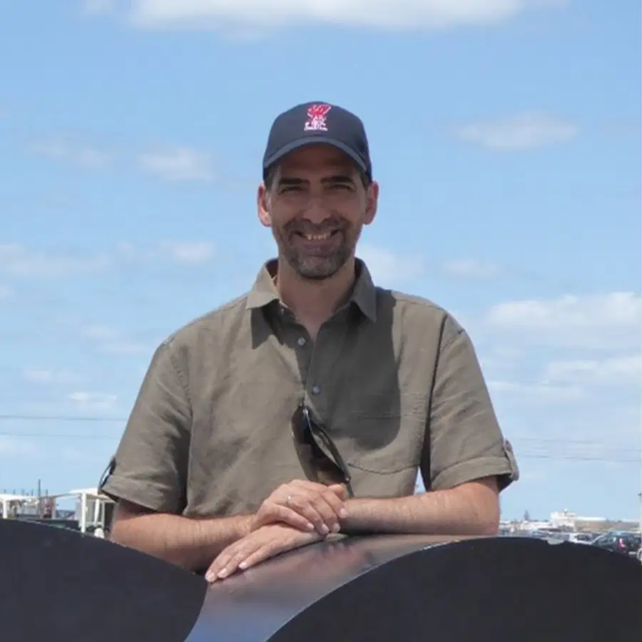 A smiling man wearing a dark baseball cap and khaki shirt stands outdoors with his hands resting on a black object. The sky is blue with scattered clouds, and boats are visible in the background.