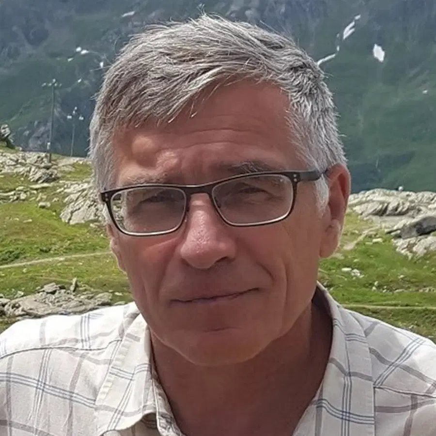 An older man with grey hair and glasses, wearing a checked shirt, stands outdoors in a mountainous area with green hills and patches of snow in the background.