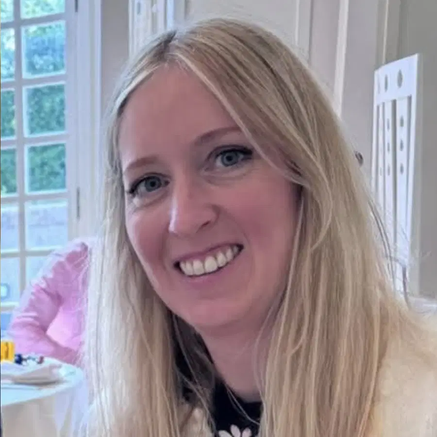 A woman with long blonde hair smiles at the camera indoors. She wears a black top and is in a well-lit room with a large window and dining table in the background.