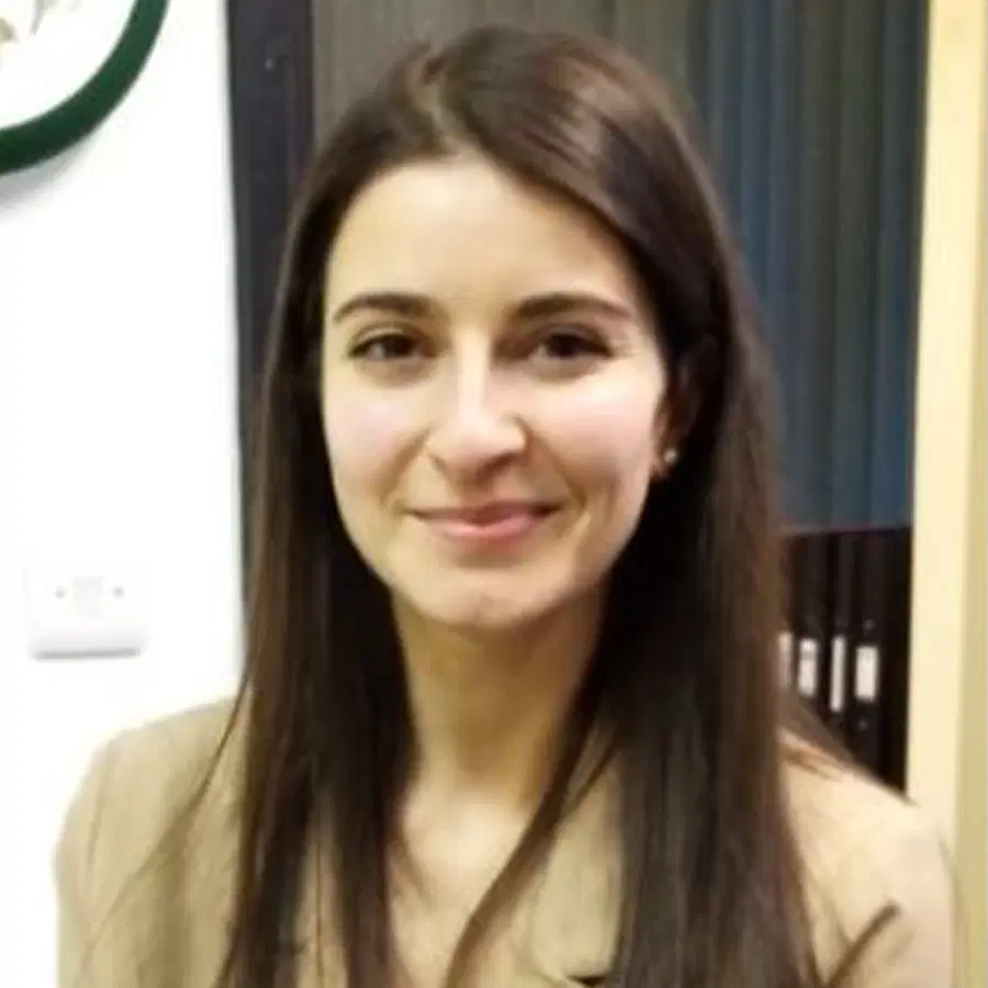 A woman with long brown hair, wearing a beige blazer, stands indoors, smiling at the camera. The background shows part of a wall, some shelves, and a green circle on the wall.