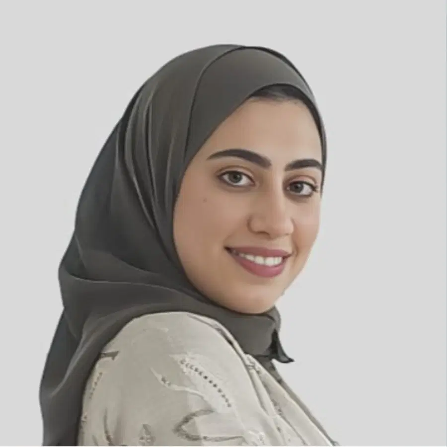 A woman wearing a grey hijab and light patterned top smiles at the camera against a plain light background.