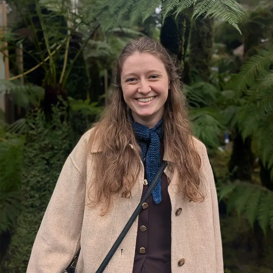 A woman with long, wavy hair smiles whilst standing outdoors among lush green ferns. She is wearing a beige coat and a blue scarf.