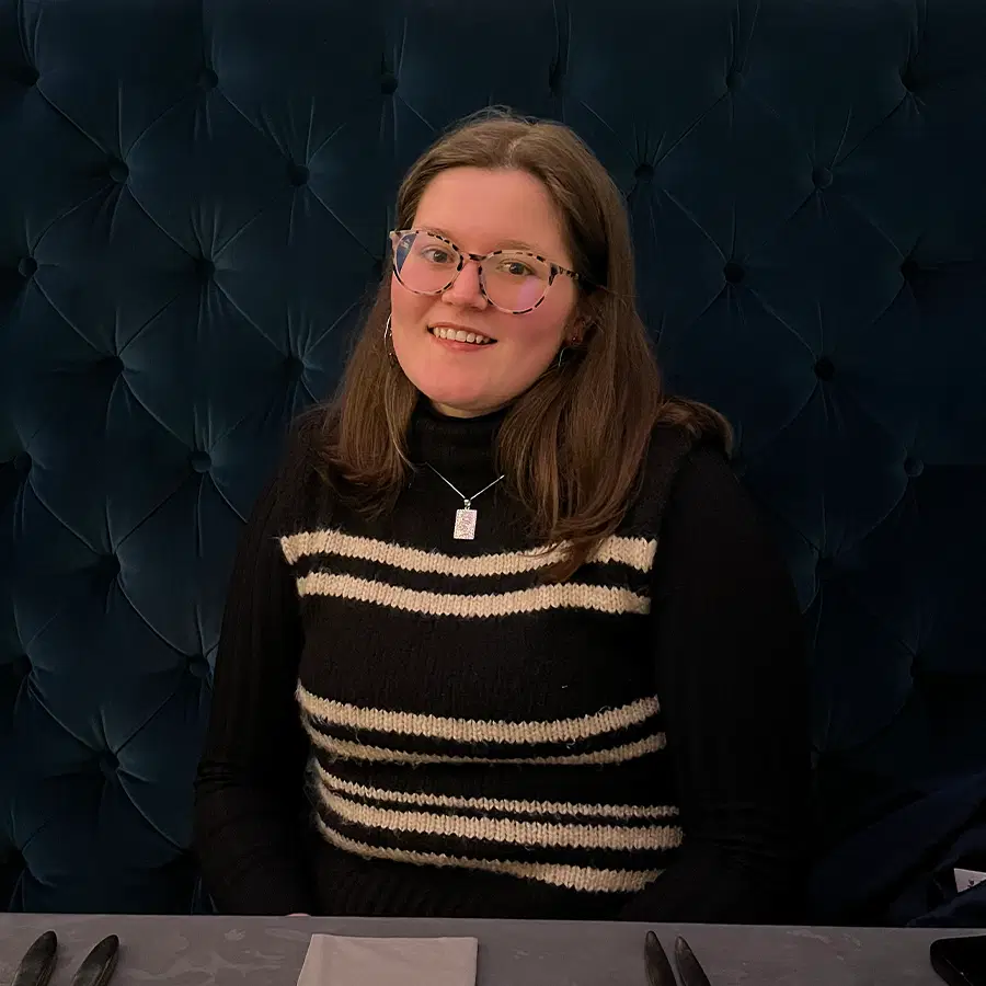 A smiling person with glasses and long brown hair sits at a table, wearing a black polo neck with a black and white striped waistcoat. The background is a deep blue, tufted booth.