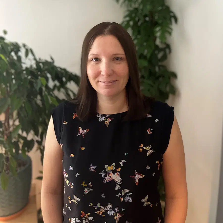 A woman with straight brown hair stands indoors, smiling slightly. She is wearing a black shirt with colourful butterfly prints. There are green houseplants in the background.
