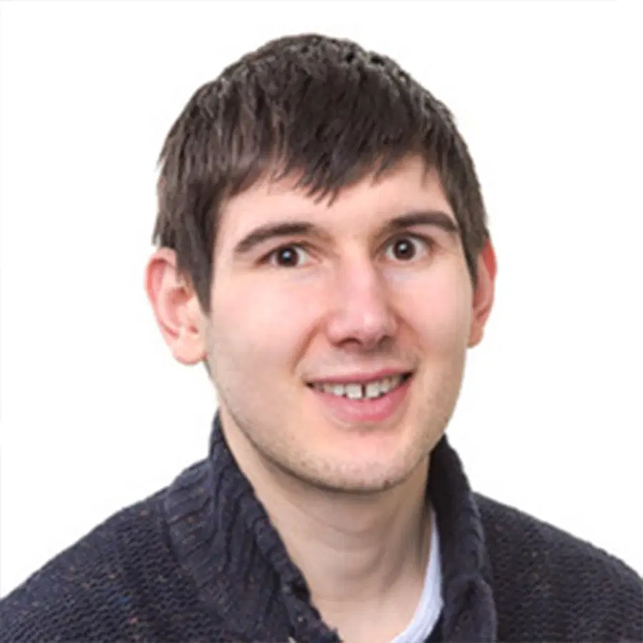 A young man with short, dark brown hair, wearing a dark textured jumper over a white shirt, smiles at the camera against a plain white background.