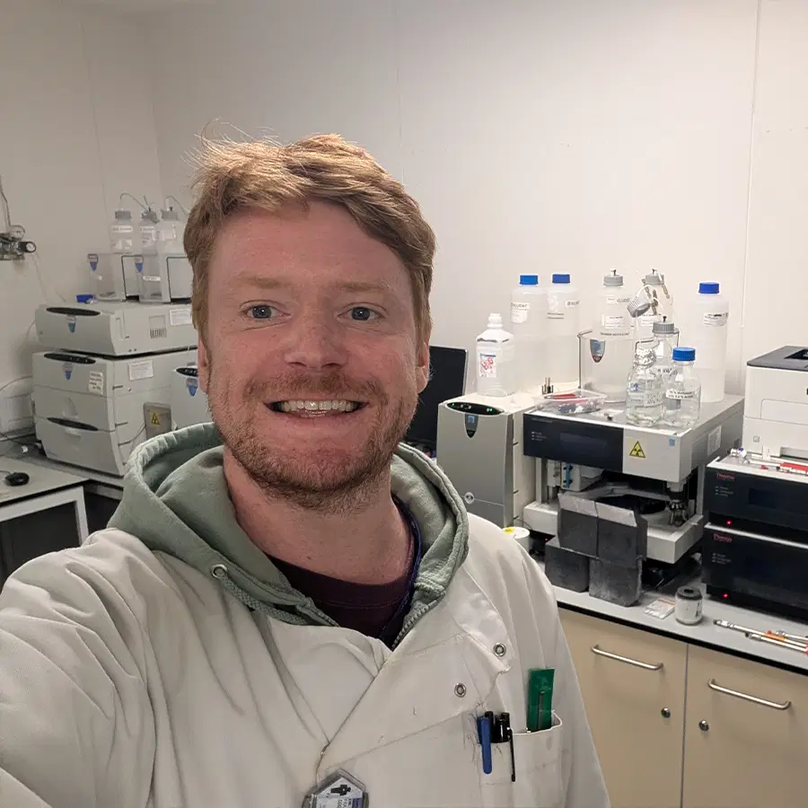 A person in a white lab coat smiles for a selfie in a laboratory, surrounded by scientific equipment, bottles, and machines on worktops and shelves.