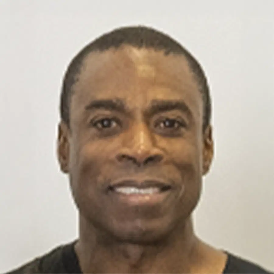 A man with short dark hair smiles at the camera against a plain light background. He is wearing a dark shirt.
