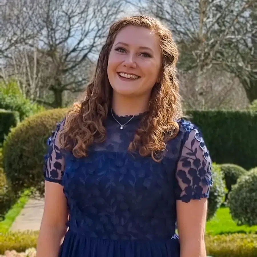 A woman with curly red hair, wearing a navy blue lace dress, stands smiling in a sunlit garden with trees and manicured hedges in the background.