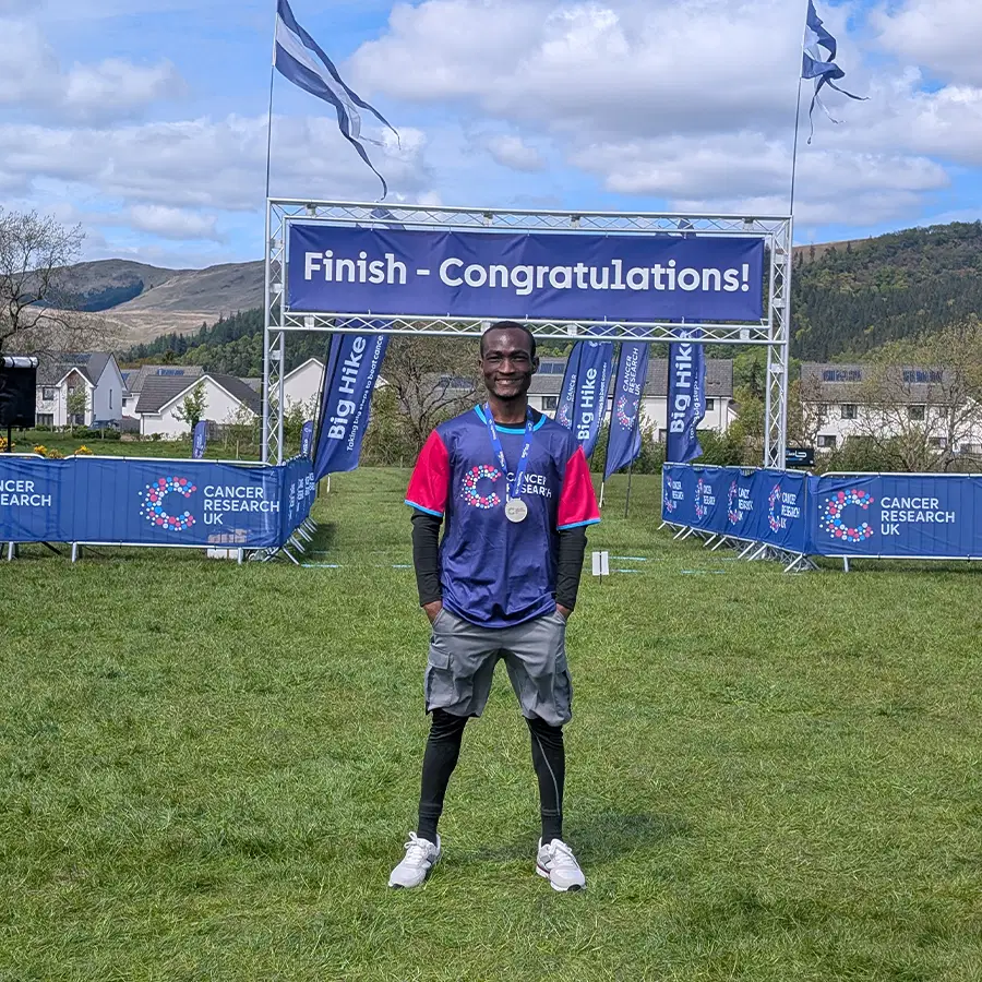 A smiling runner wearing a medal poses in front of a Finish - Congratulations! sign at an outdoor event, with Cancer Research UK banners and flags in the background.