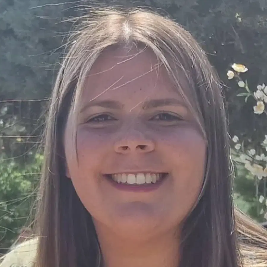 A person with long, straight hair smiling outdoors, with green foliage and small white flowers in the blurred background.