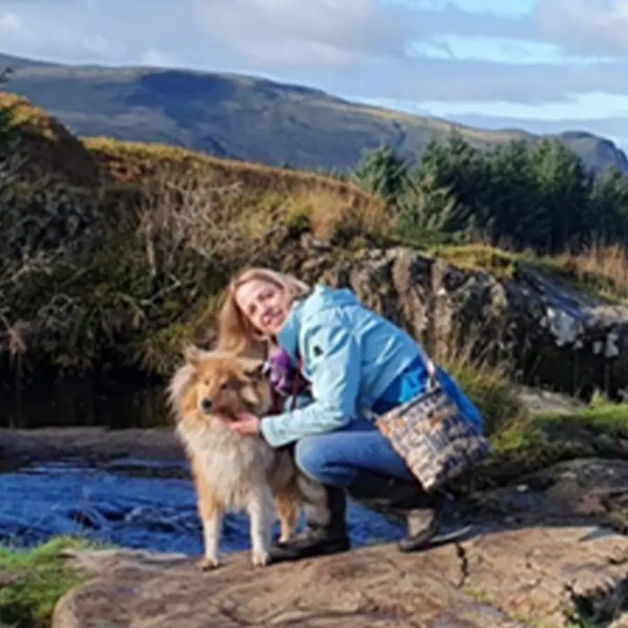 A person in a blue jacket kneels beside a brown and white dog on a rock. Behind them are rolling hills, trees, and a stream under a partly cloudy sky.