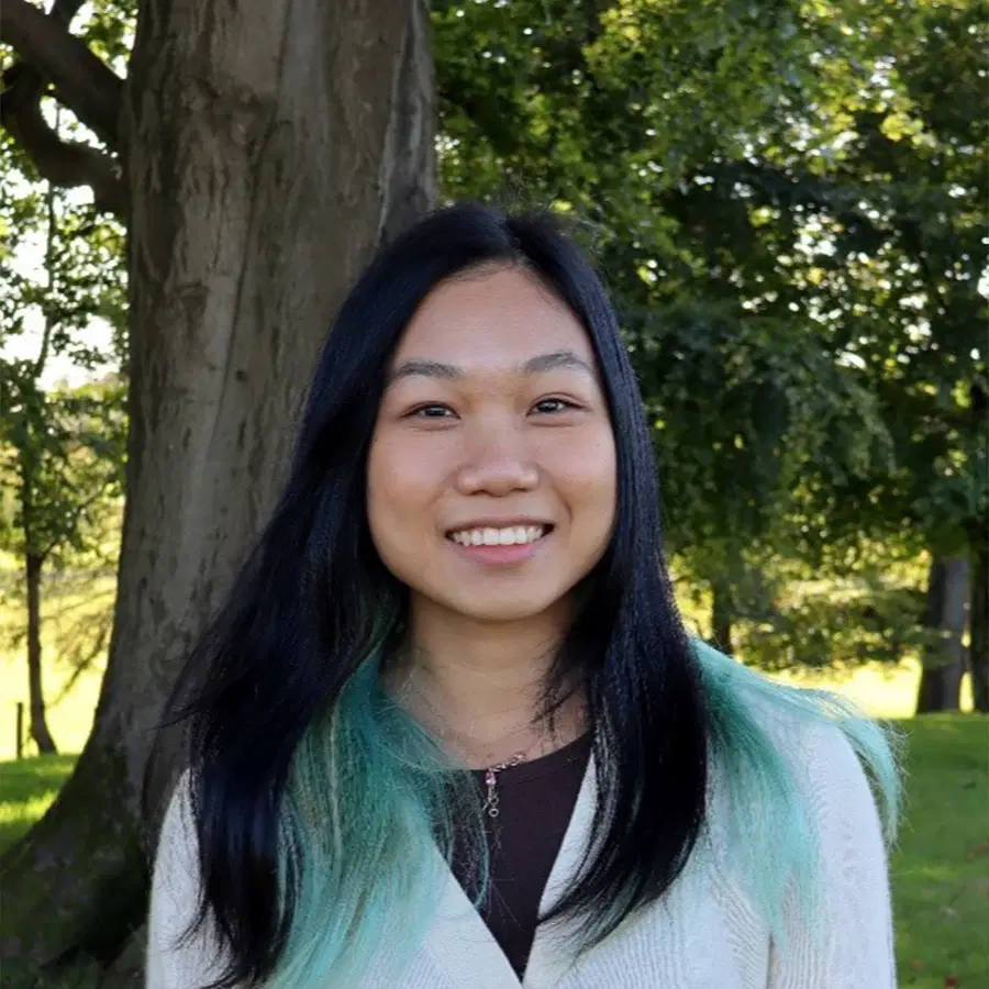 A person with long black hair and green tips smiles outside, standing in front of a tree and lush green foliage on a sunny day.