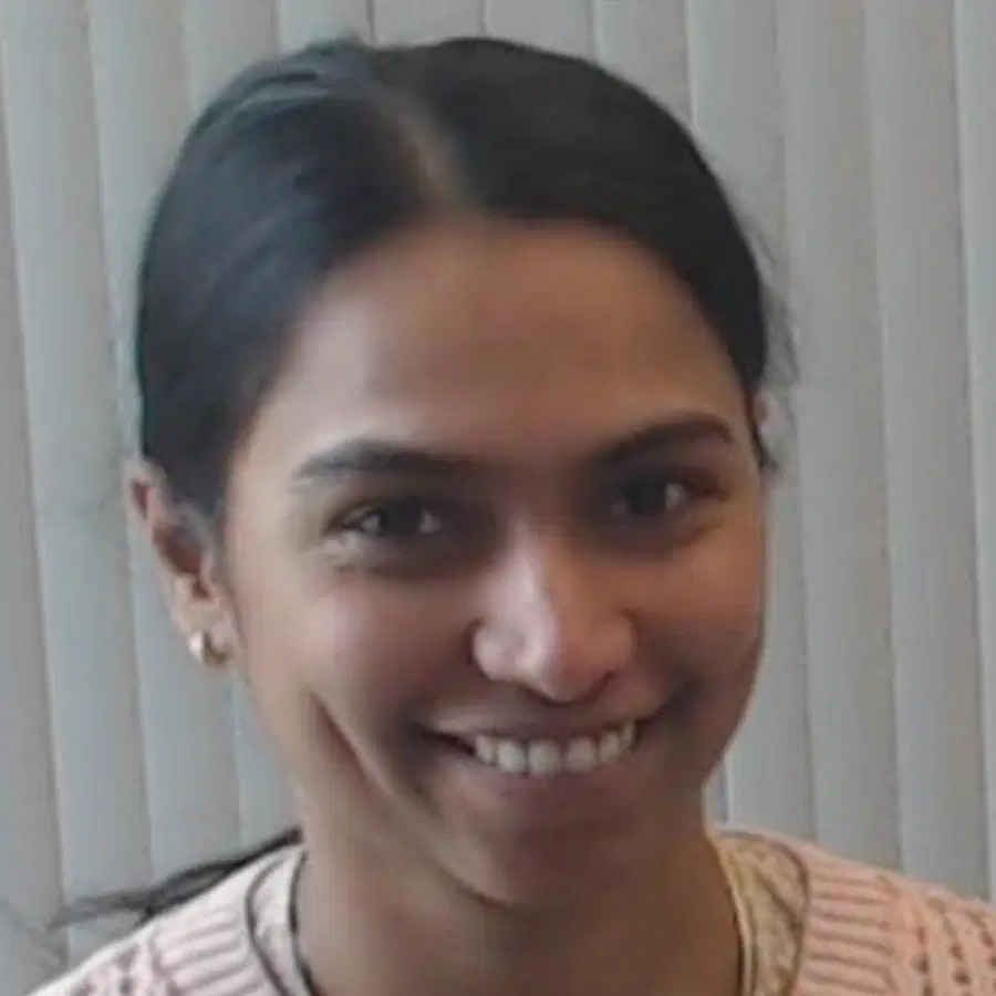 A person with long dark hair tied back smiles at the camera, standing in front of vertical blinds and wearing a light-coloured top.