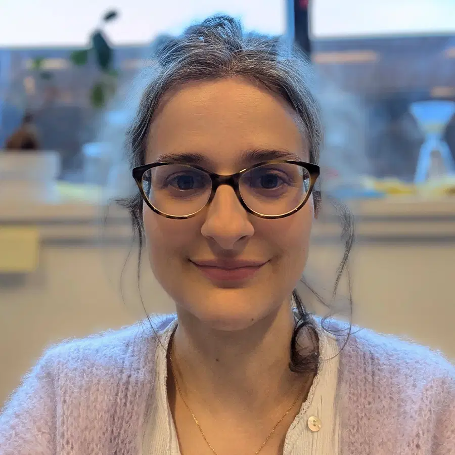 Woman with long brown hair in a loose bun, wearing glasses and a lavender cardigan, smiles at the camera in a softly lit indoor setting with a blurred background.