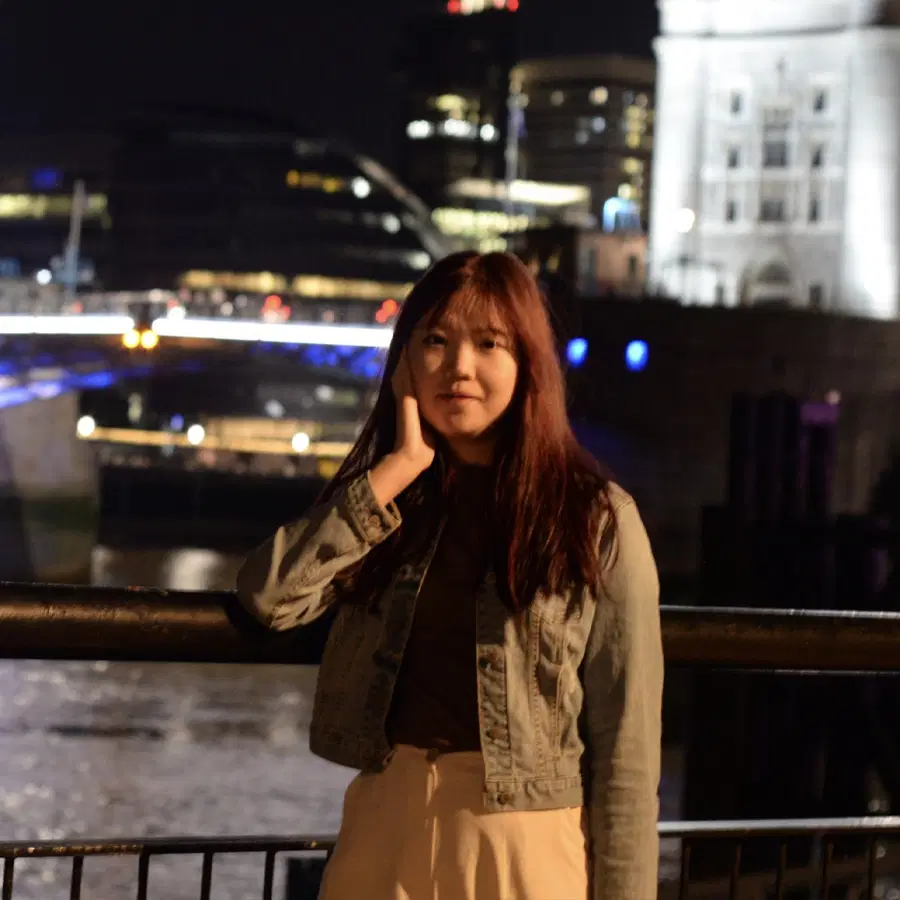 A young woman with long brown hair stands by a river railing at night, touching her face, with illuminated modern buildings and a bridge in the background.