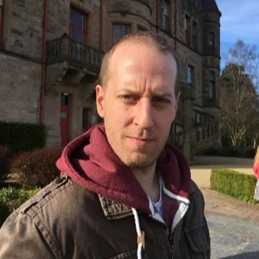 A man in a brown jacket and maroon hoodie stands outside a large stone building, facing the camera on a sunny day. Some greenery is visible in the background.