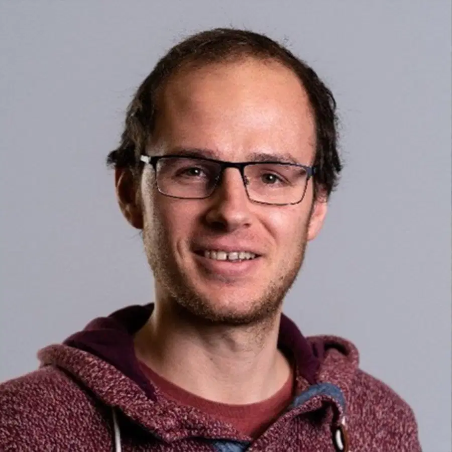 A man with short brown hair, glasses, and a maroon hoodie smiles at the camera against a plain grey background.
