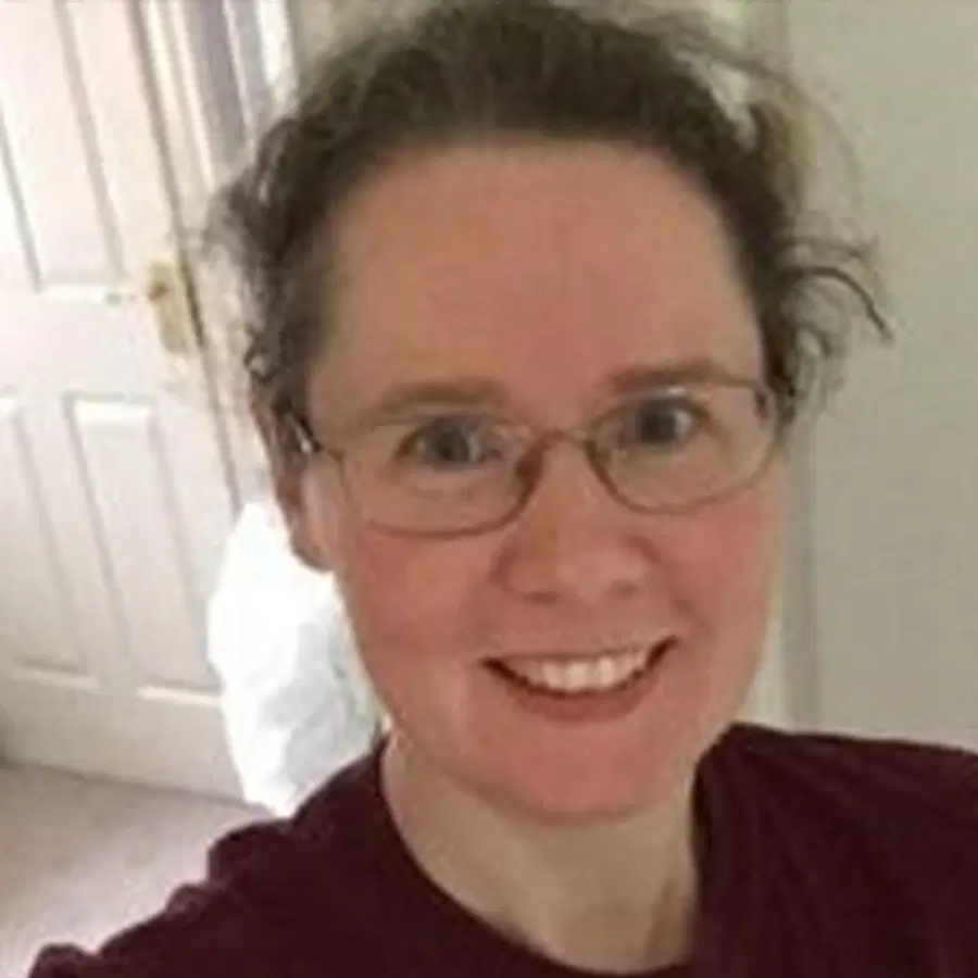 A person with glasses and curly brown hair pulled back smiles at the camera whilst standing indoors near a white door.
