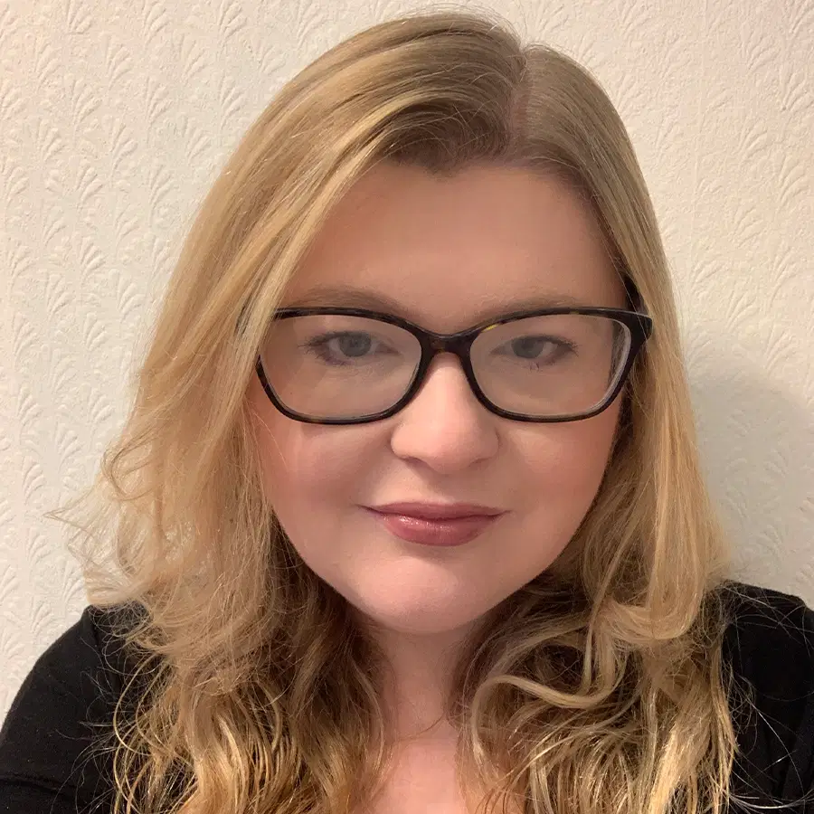 A person with long blonde hair and glasses is smiling slightly, posing indoors in front of a light patterned wall, wearing a black top.