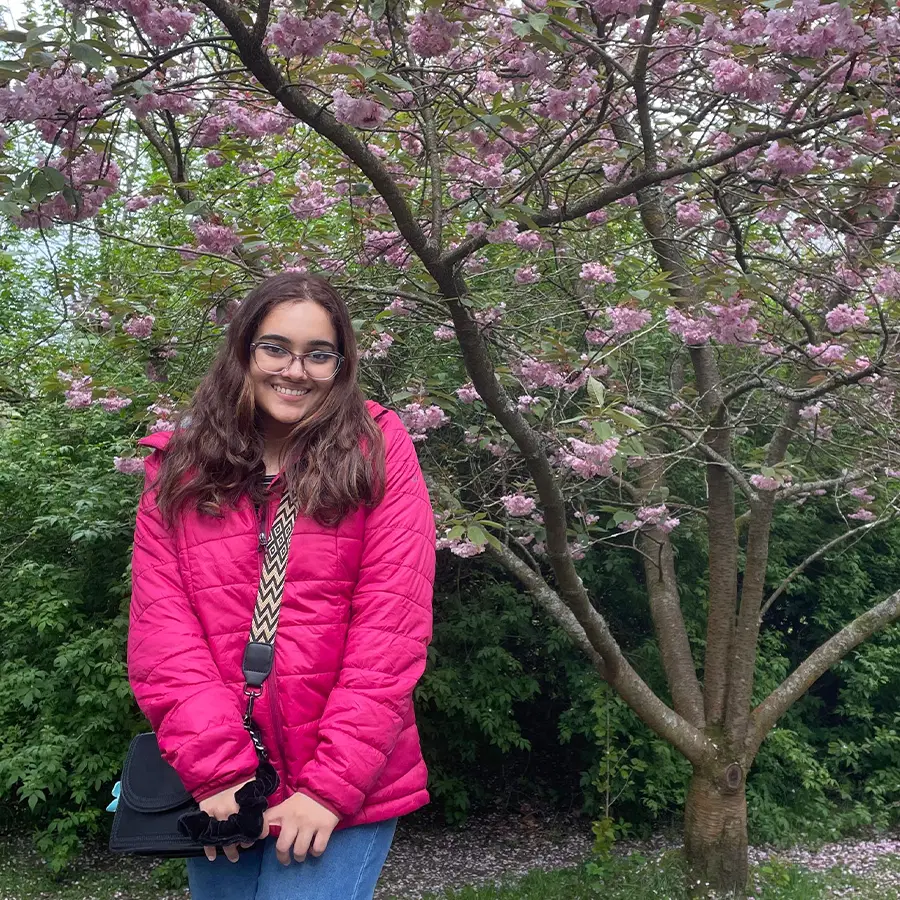 A smiling person wearing a bright pink jacket stands in front of a blossoming tree with pink flowers, surrounded by lush green foliage.