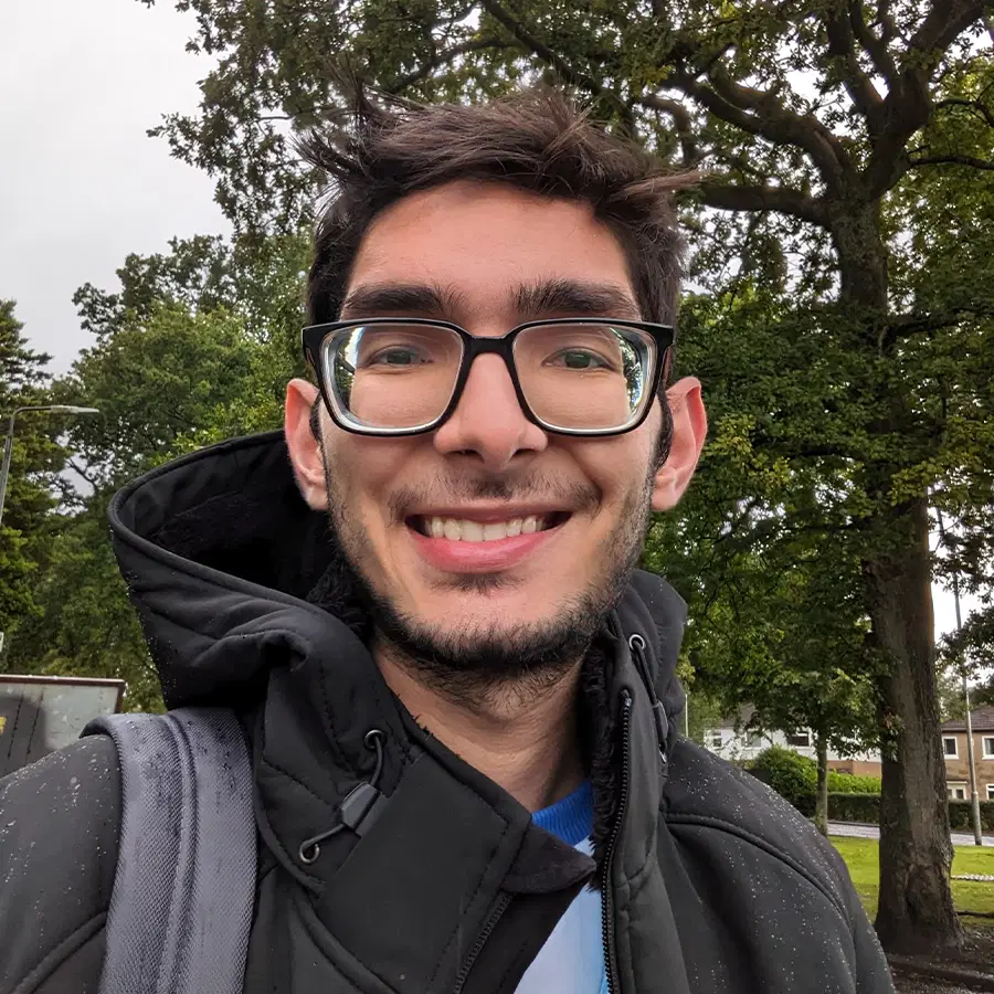 A person with glasses and a beard smiles at the camera outdoors, wearing a black jacket and carrying a grey rucksack. Green trees and houses are visible in the background.