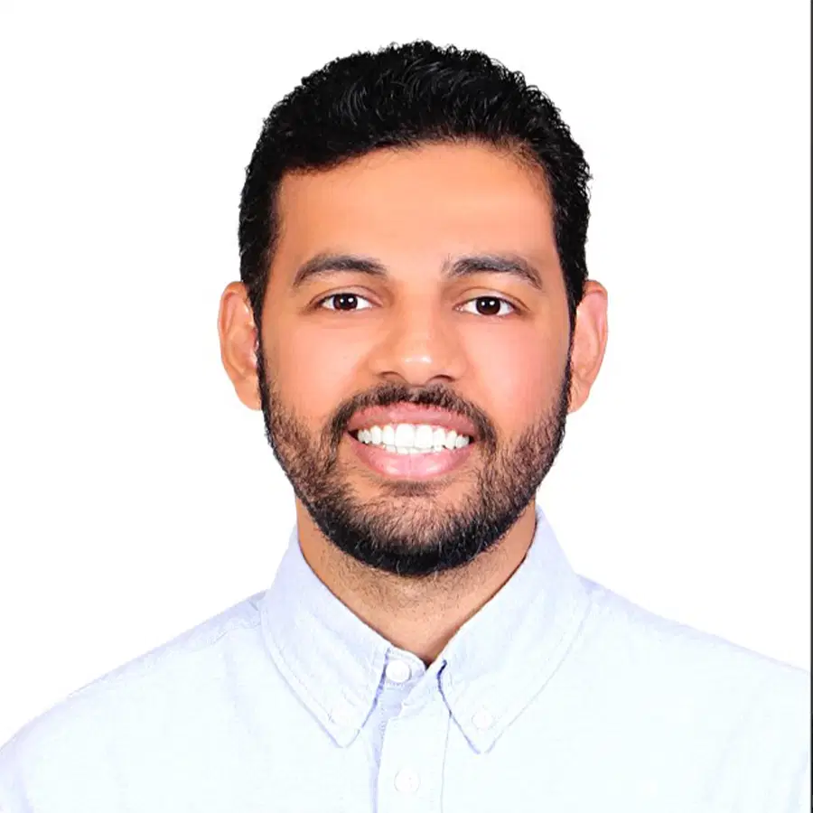 A man with short dark hair, a beard, and moustache, wearing a light blue collared shirt, smiling in front of a plain white background.