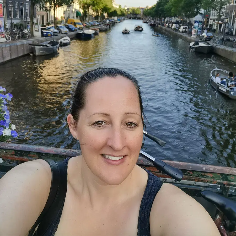 A woman smiles for a selfie on a bridge over a canal lined with boats and trees in a city, likely during the evening.