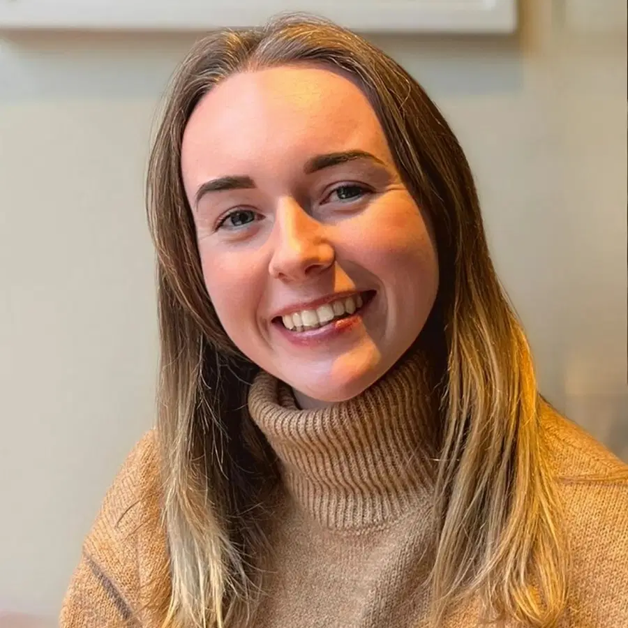 A young woman with long, straight, light brown hair smiles whilst wearing a tan polo-neck jumper, sitting indoors with natural light on her face.